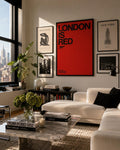 May include: A stylish living room featuring framed artwork, a white sofa, and a marble coffee table. The art includes a red poster with the text "LONDON IS RED" and a black and white photograph of a building. A large plant and books are also present.