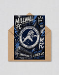 May include: A greeting card with the Millwall FC crest, a lion rampant within a circular design, and the text "MILLWALL FOOTBALL CLUB" and "1885". The card has a distressed aesthetic with blue and white details, and the slogan "NO ONE LIKES US WE DON'T CARE".